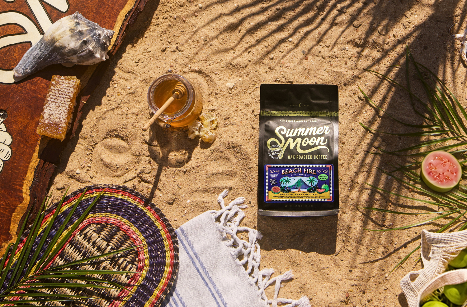 Summer Moon Beach Fire coffee bag lying on sand, surrounded by honeycomb, a jar of honey, a woven basket, seashell, palm leaves, and sliced guava. 