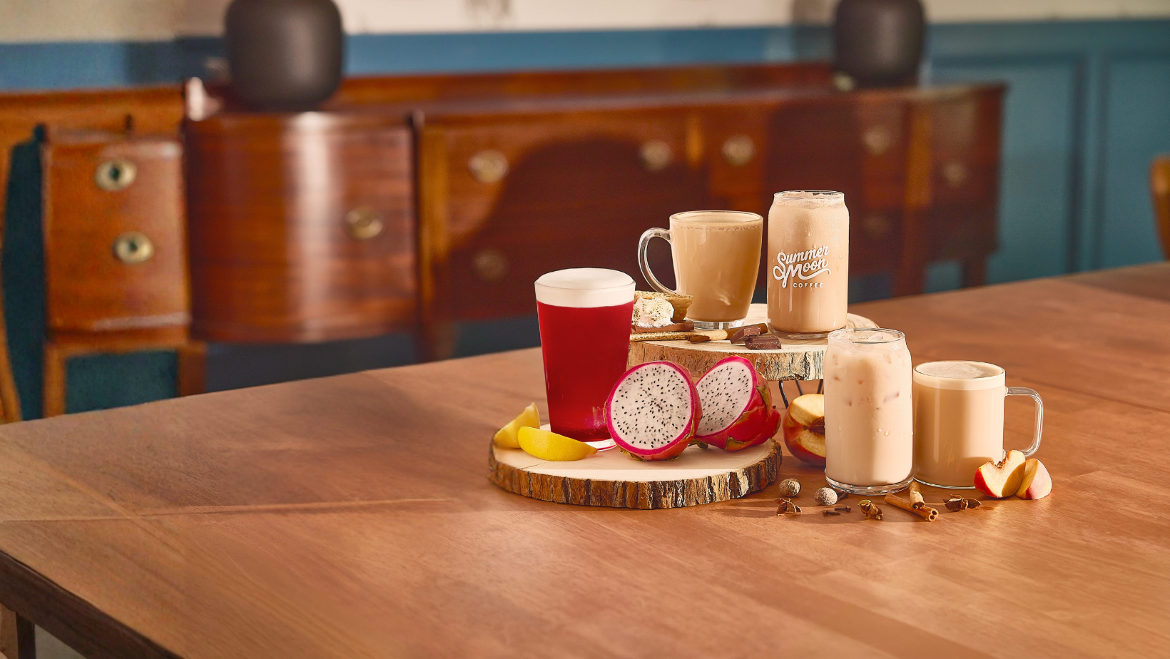 A styled tabletop arrangement of assorted Summer Moon Coffee beverages on a wooden table, including a red drink with foam, two tan coffee drinks, two frothy milk drinks, sliced dragon fruit, and fruit pieces, with a blurred indoor room in the background.