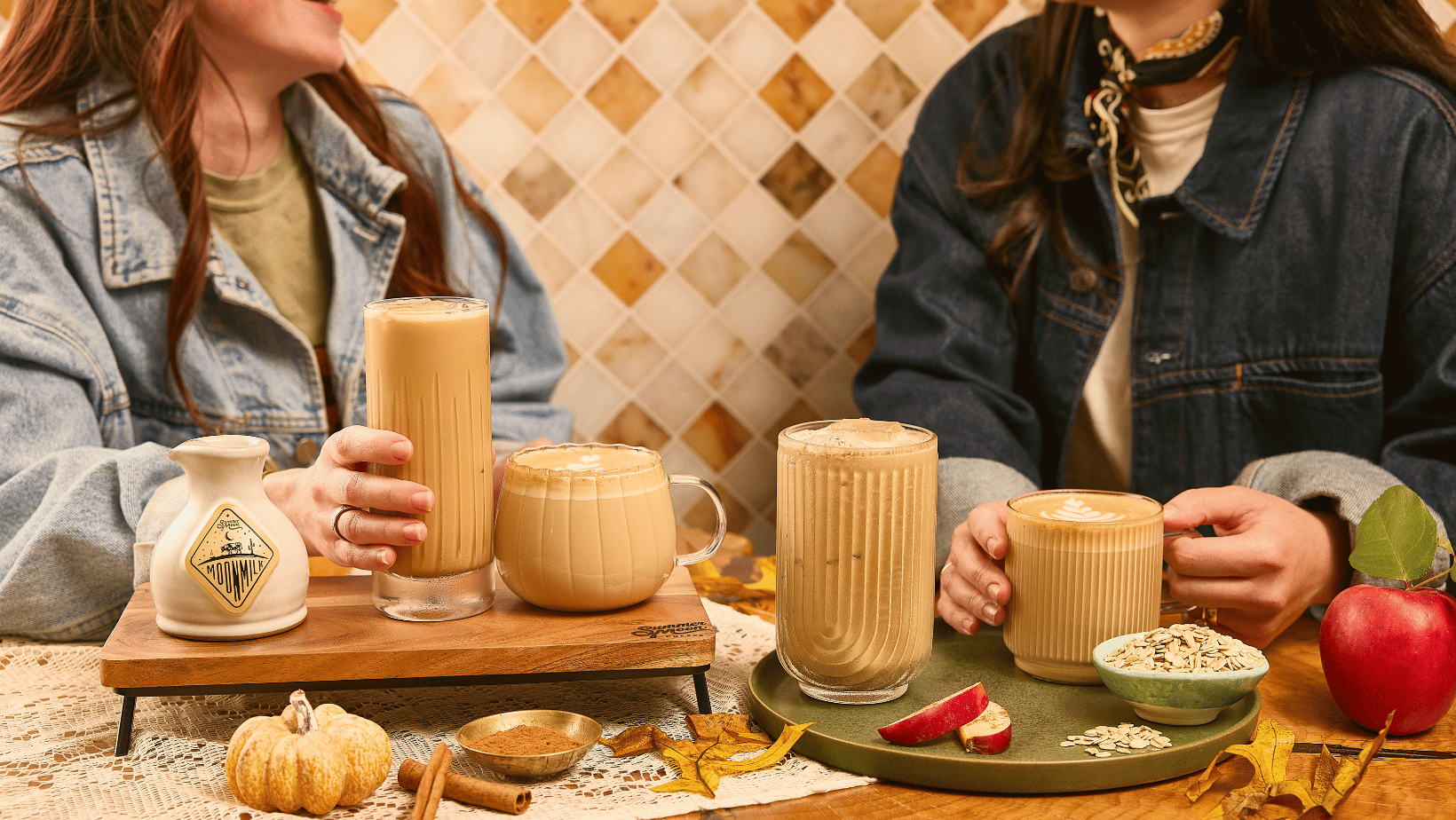 Two women in denim jackets sharing iced and hot Summer Moon lattes at a table with a Moon Milk pitcher, cinnamon, apple slices, oats, and a small pumpkin.