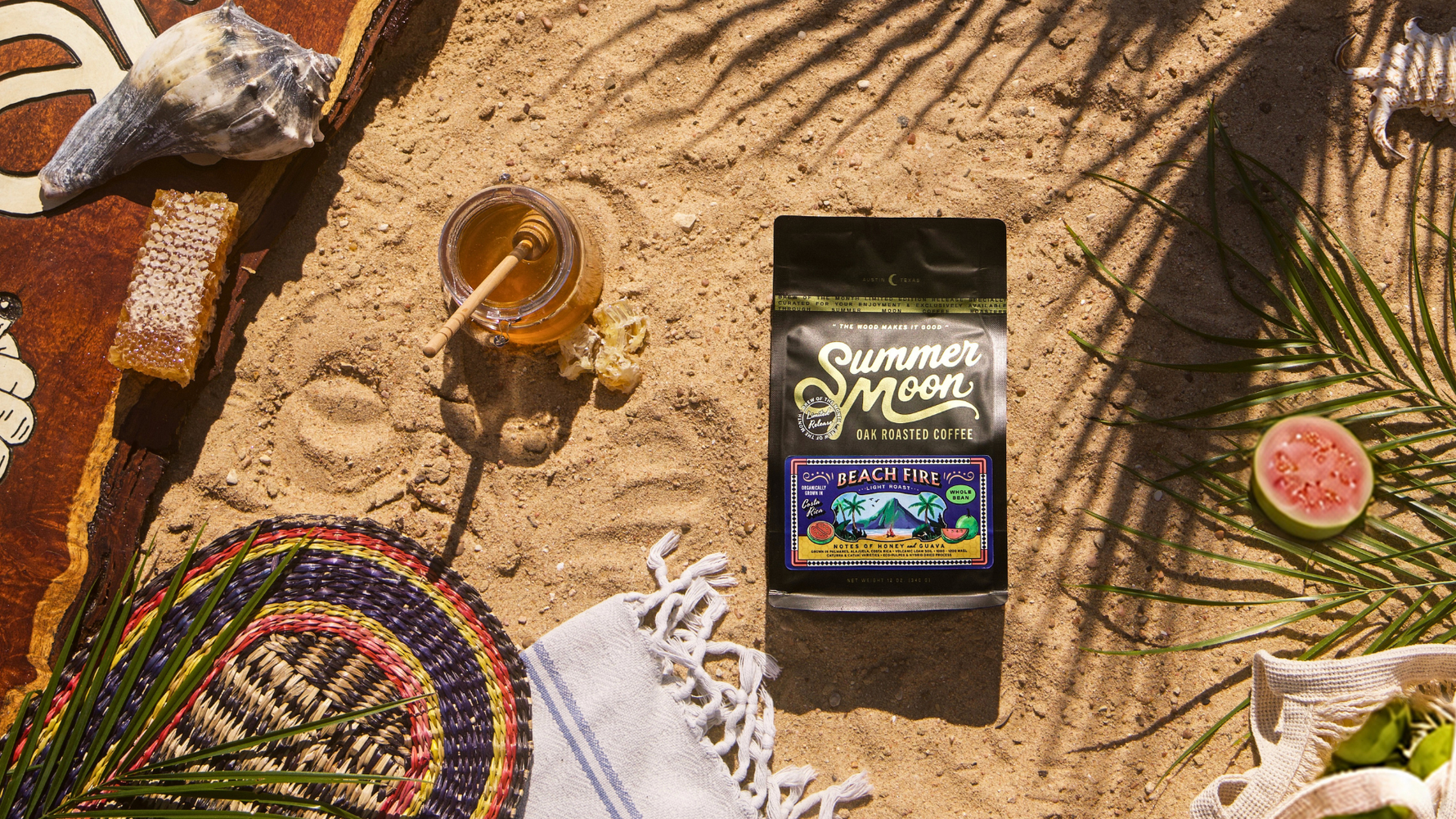 Summer Moon Beach Fire coffee bag lying on sand, surrounded by honeycomb, a jar of honey, a woven basket, seashell, palm leaves, and sliced guava.