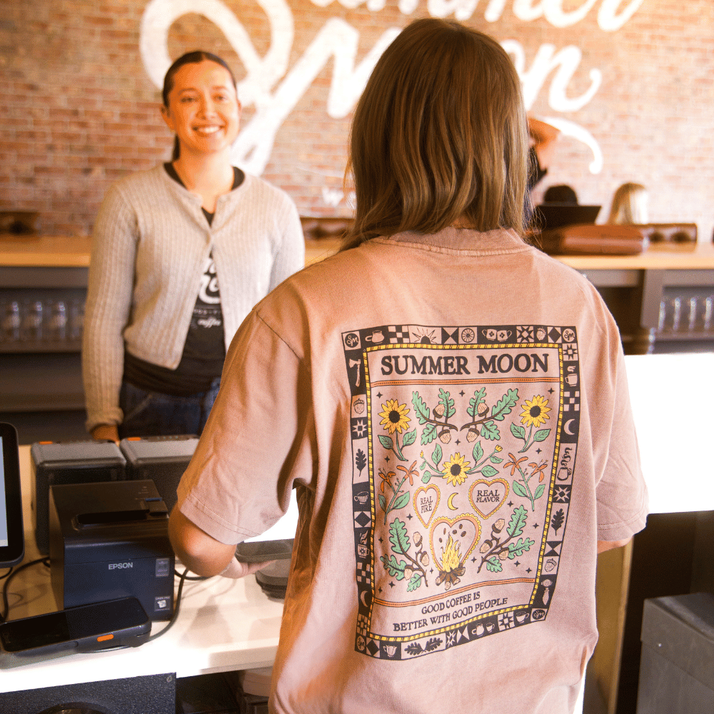 A person viewed from behind wearing a light pink graphic T-shirt with a large 'Summer Moon' sunflower and heart design on the back, standing at a coffee counter with a smiling barista in a brick-walled cafe.