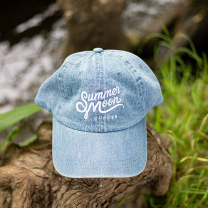 Light wash denim unstructured dad hat rests on a tree stump. 