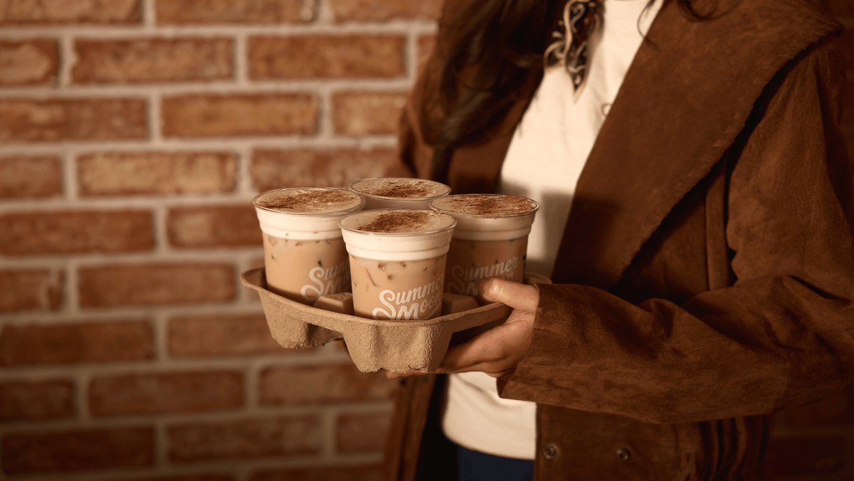 A person in a brown suede jacket and white shirt holds a tray with four iced chai from Summer Moon Coffee in front of a warm brick wall.
