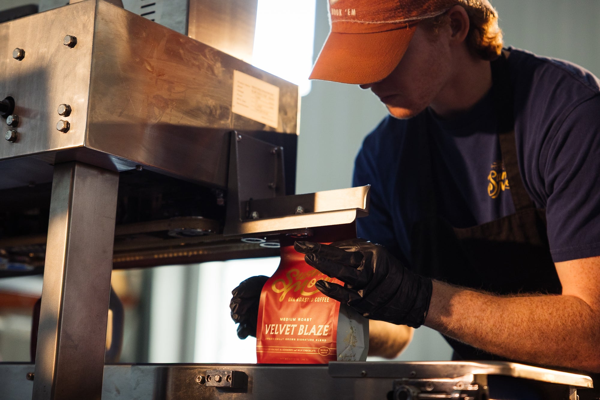 A person wearing a navy blue t-shirt, brown apron, and orange cap uses a large metal machine to seal a red coffee bag labeled 'VELVET BLAZE'.